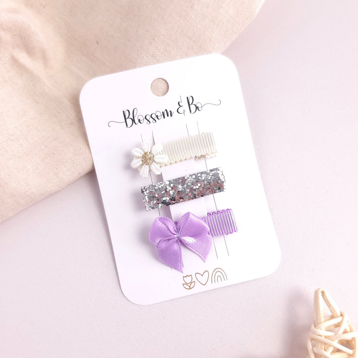 A set of three hair clips on a white display card, featuring a white flower clip, a silver glitter clip and a lavender ribbon bow clip. The card is branded 'Blossom & Bo' and is styled on a light pink background with soft beige fabric and a small decorative woven object.