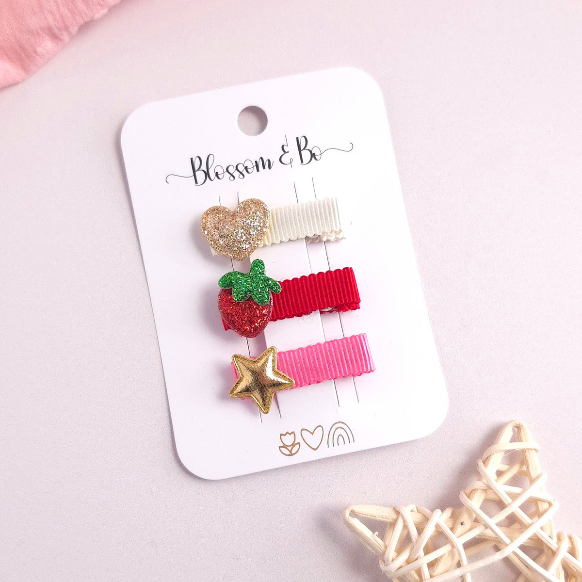 Three children's hair clips on a white display card labeled 'Blossom & Bo,' photographed on a pale pink background with decorative props.