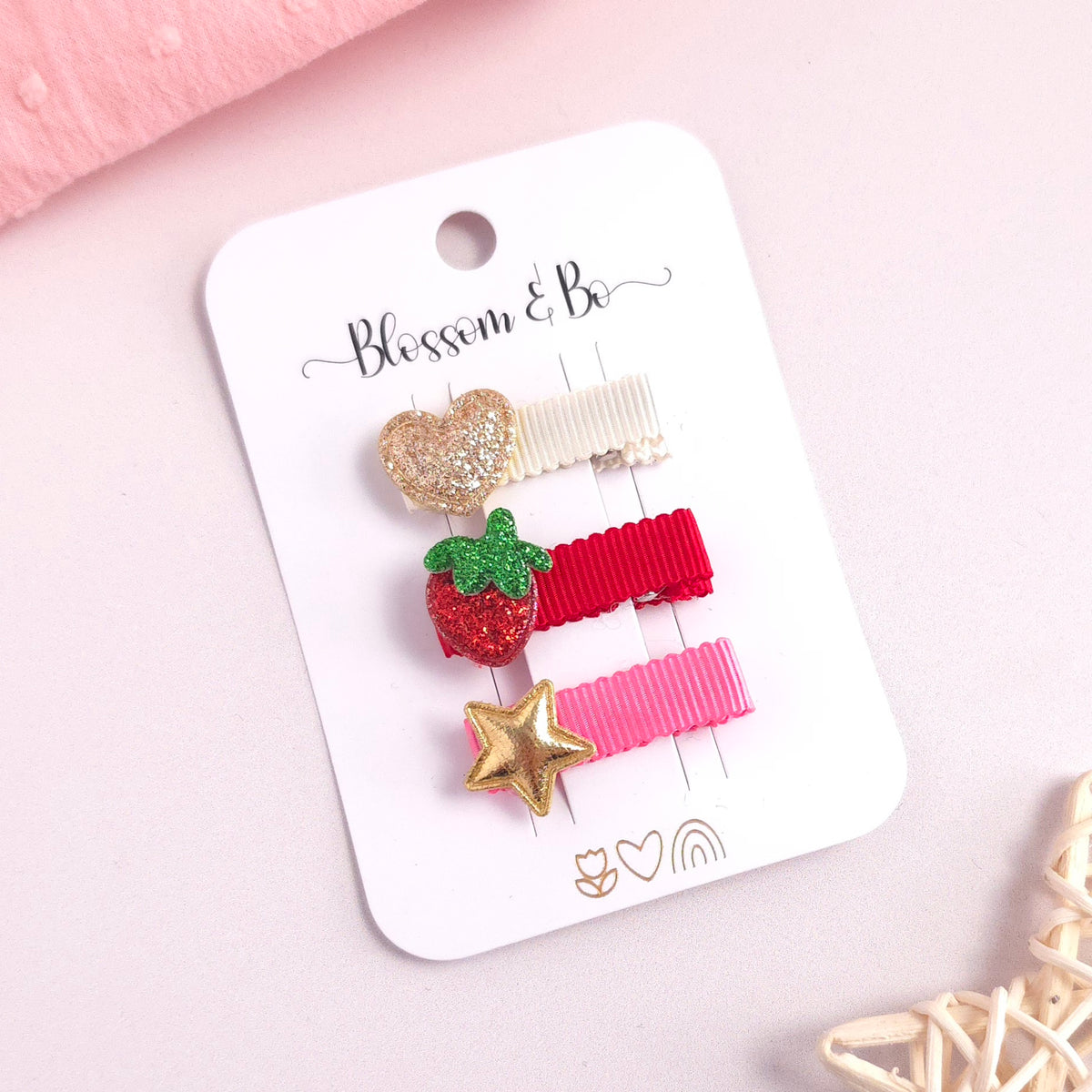 Three kids' hair clips on a white card from the brand Blossom & Bo, styled on a light pink background with decorative props.
