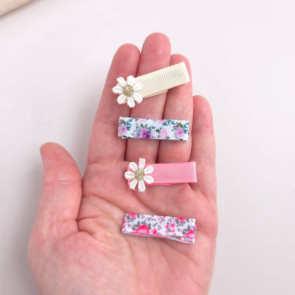 Four hair clips held in a person's hand, including two floral ribbon clips, one cream ribbon clip with a white flower accent, and one pink ribbon clip with a white flower accent.