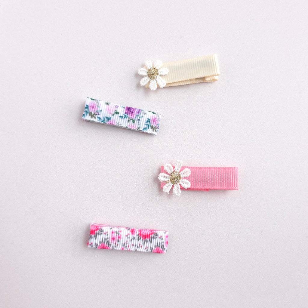 Four small hair clips arranged on a light pink background. Two clips are floral with pink, purple, and white ribbon, and two are solid pastel pink and cream with white daisy flower embellishments.