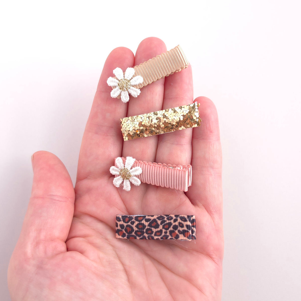A hand holds four hair clips against a plain light background. The clips include a ribbed tan band, a glittery gold band, a ribbed pink band, a white flower clip with a gold center, and a leopard-print band.