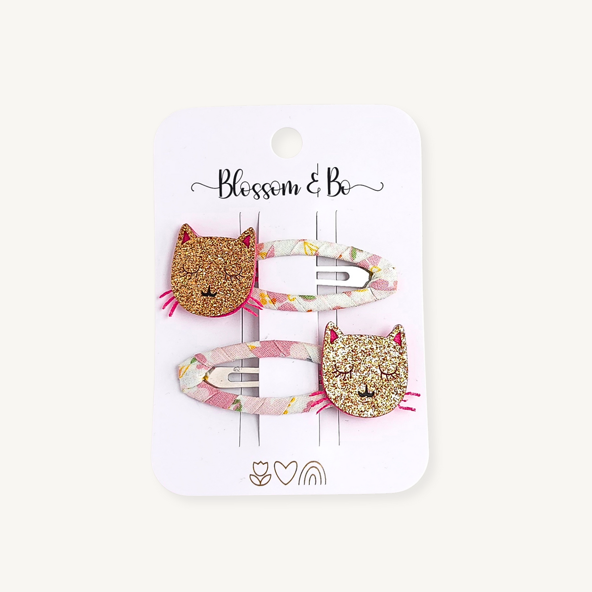 Two hair clips on a white card backing. The clips have glittery gold cat faces with pink ears and noses, attached to pastel pink, yellow, and white patterned bands.