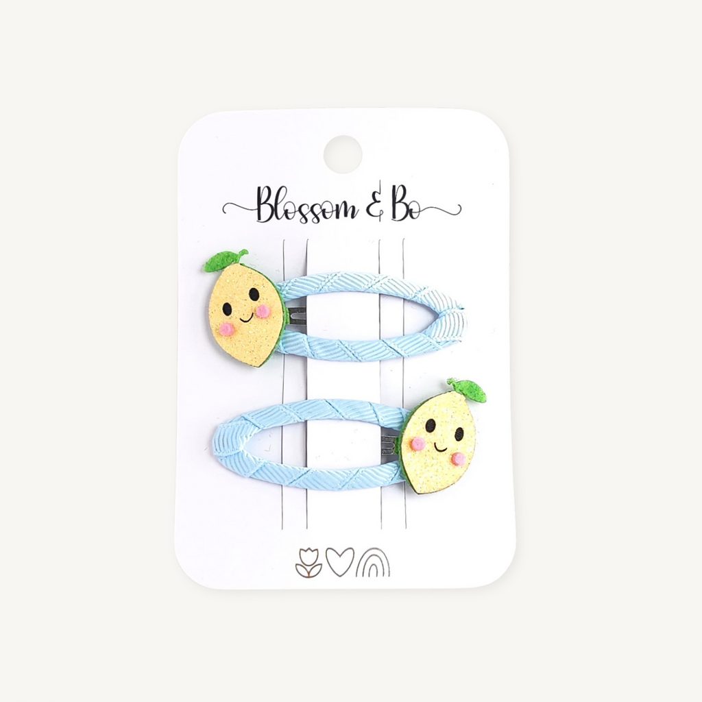 Set of two blue fabric covered snap hair clips featuring smiling lemon embellishments on a Blossom & Bo display card.