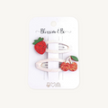 A set of two cream-colored woven hair clips decorated with a red glittery strawberry and a pair of red glittery cherries with green leaves, presented on a white backing card labeled 'Blossom & Bo'.