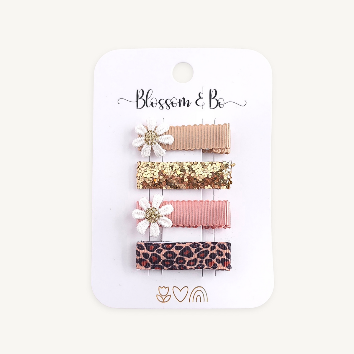 Four hair clips on a white card backing with the brand name 'Blssom & Bo' printed at the top. The clips include a tan ribbed clip with a white flower, a gold sequin clip, a pink ribbed clip, and a leopard print clip.