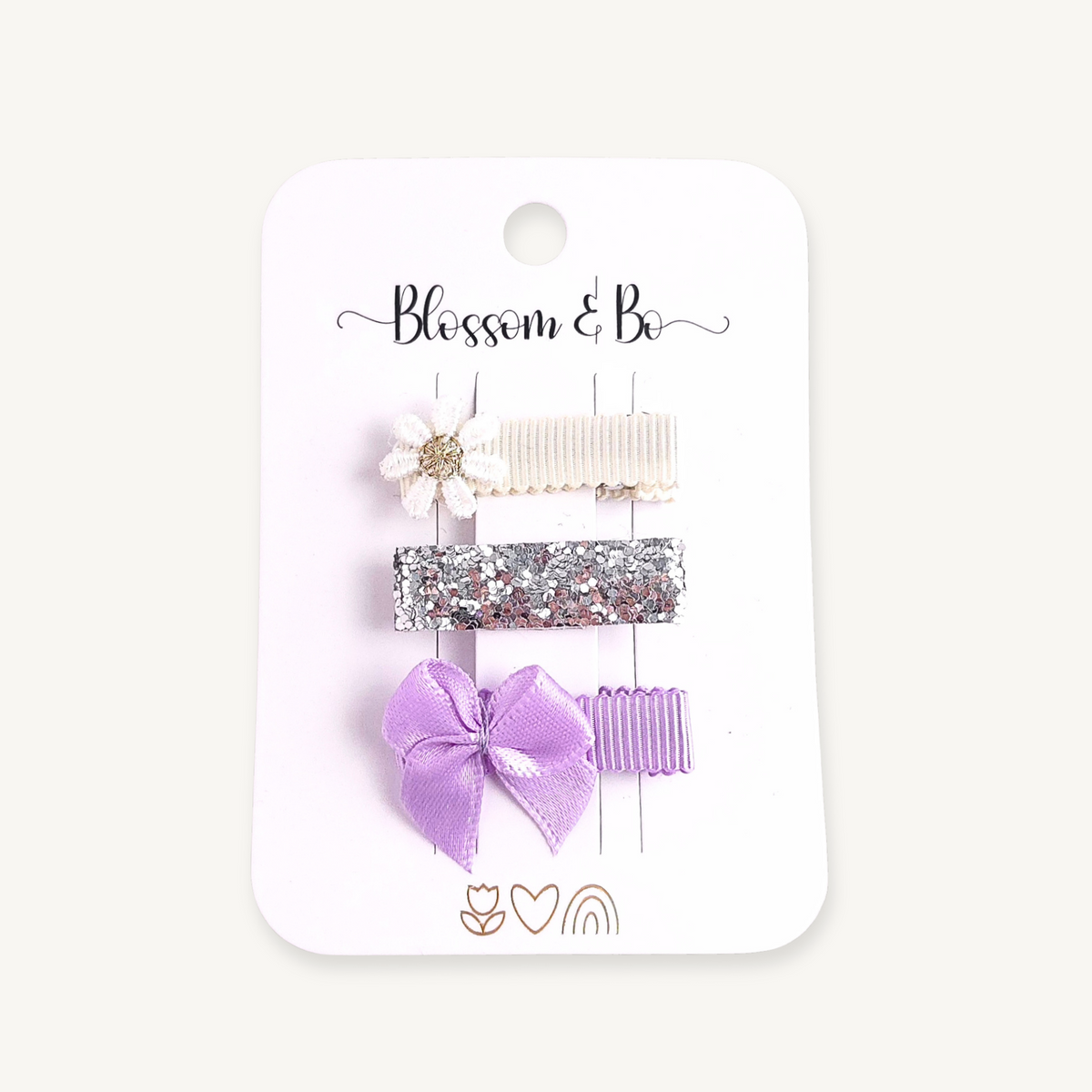 Three hair clips on a white display card with the brand name 'Blossom & Bo' printed at the top. The clips include a white fabric flower with a gold centre on a cream lined clip, a silver glitter clip and a lavender satin bow clip.