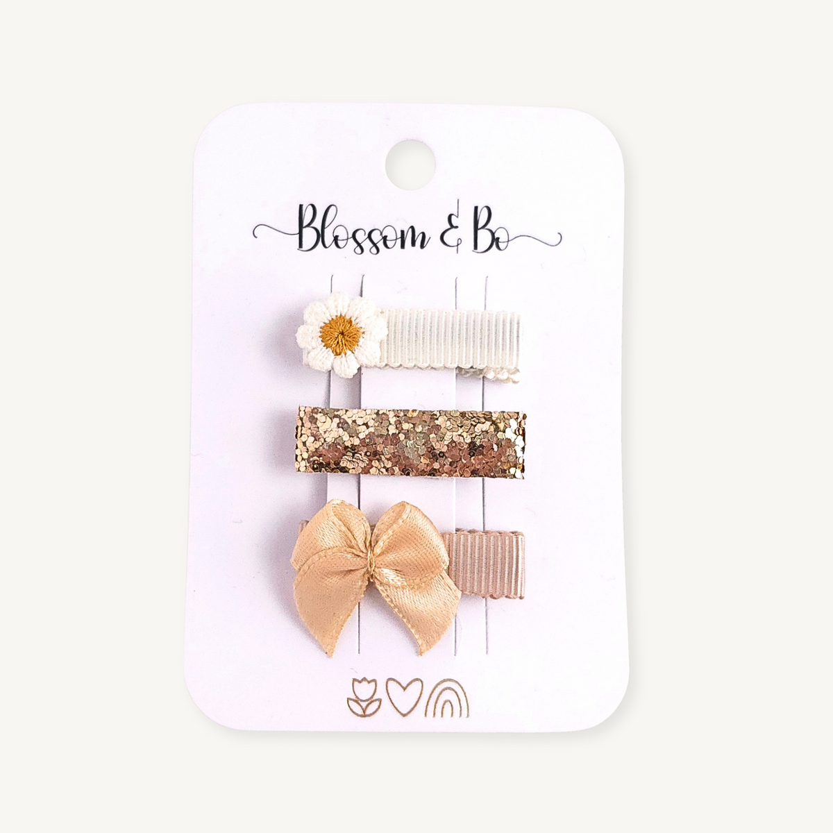 A set of three hair clips on a white card backing printed with the brand name 'Blossom & Be'. The clips include a white daisy, a gold glitter bar, a beige satin bow, and a beige ribbed bar.