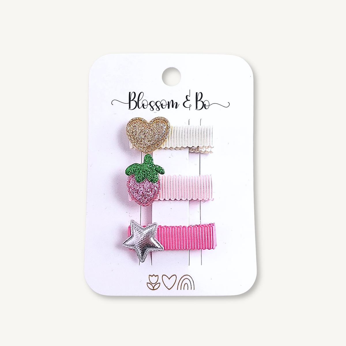 A set of three hair clips on a white backing card. The clips include a glittery gold heart, a pink strawberry with green leaves, and a silver glitter star, all attached to pink and cream ribbons.