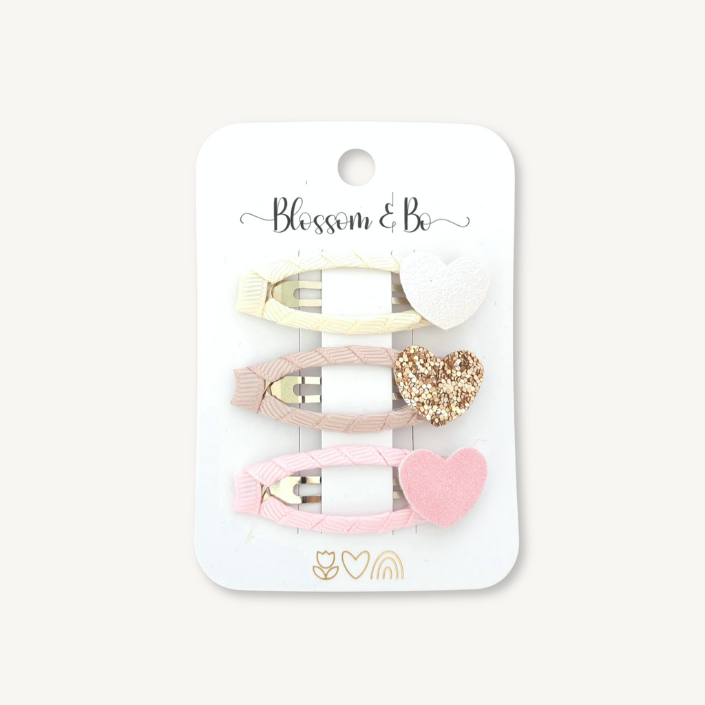 A set of three neutral hair clips on a white card backing. The clips include a cream clip with a white textured heart, a light pink clip with a glittery gold heart, and a light pink clip with a solid pink heart.