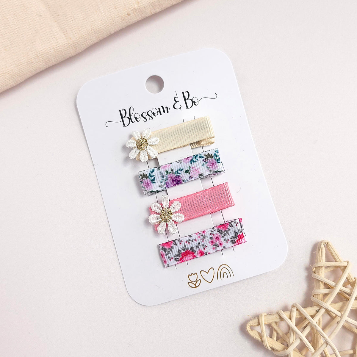 Four hair clips attached to a white card labeled 'Blossom & Bo,' photographed on a light beige surface with soft decorative props. The clips include two floral patterned bars, one cream clip, and a pink ribbed clip, with two white flower clips featuring gold centers.