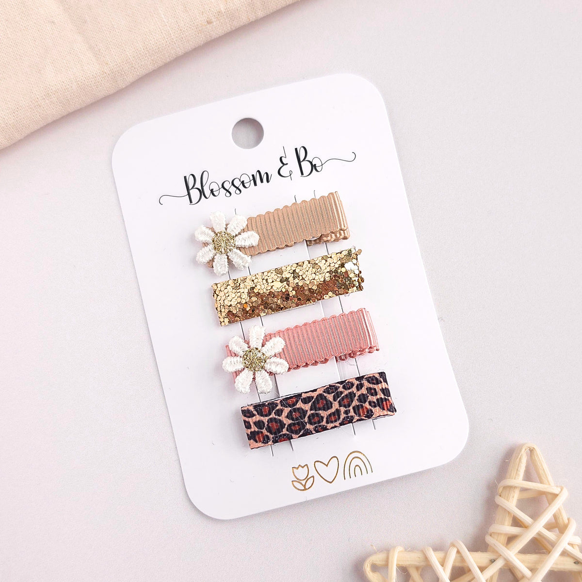 Four hair clips on a white display card labeled 'Blossom & Bo,' photographed on a light neutral surface with soft decorative props. The clips include a gold glitter bar, a white daisy with gold center, a white daisy with gold center, and a leopard-print bar.