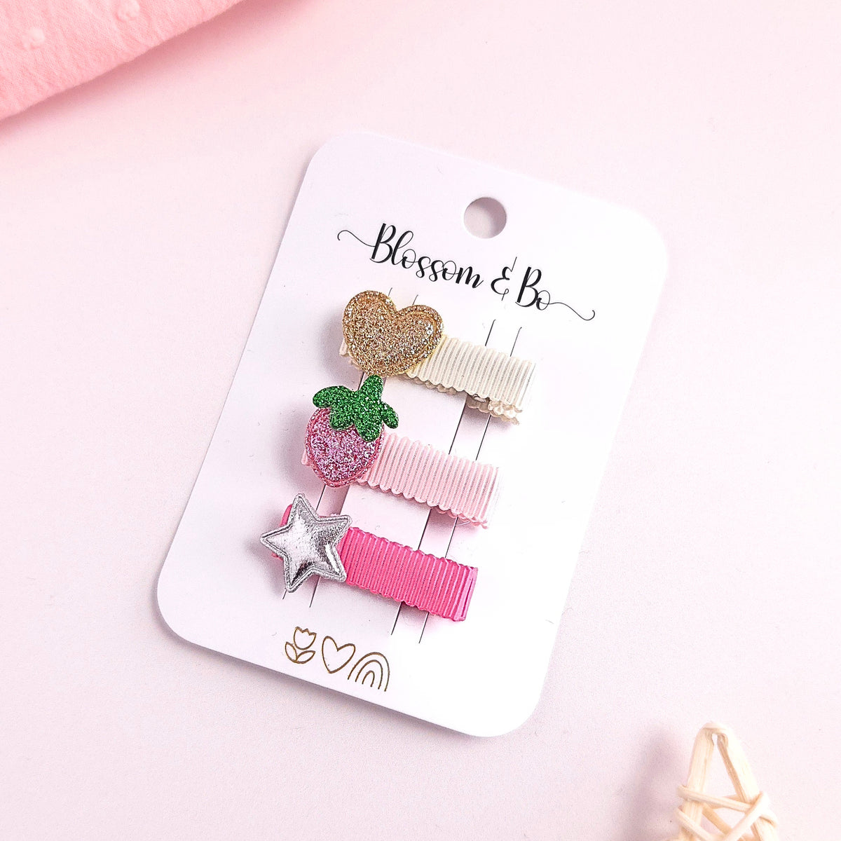 three small hair clips attached to a white card labeled 'Blessen & Bo,' photographed on a soft pink background. The clips include glittery gold, pink strawberry, silver star, and pastel pink ribbons.