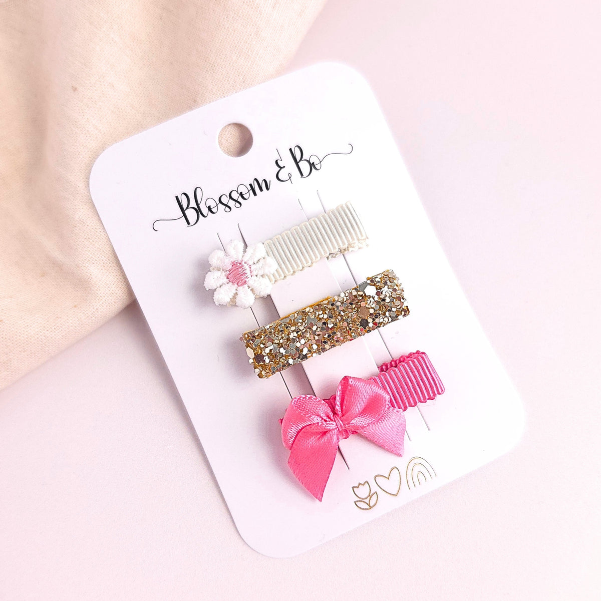 Three hair clips on a white card labeled 'Blossom & Bo' against a soft pink background. The clips include a white flower with a pink center, a cream ribbed clip, a gold glitter clip, and a pink ribbon bow clip.