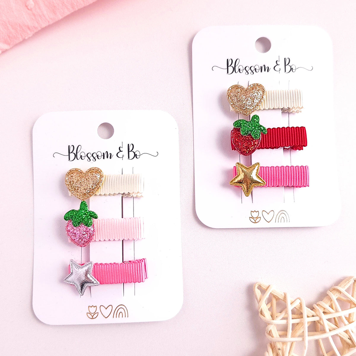 Two white carded hair clips displayed on a light pink background. The left card holds three clips: a glittery gold heart, a pink strawberry with green leaves, and a silver star.