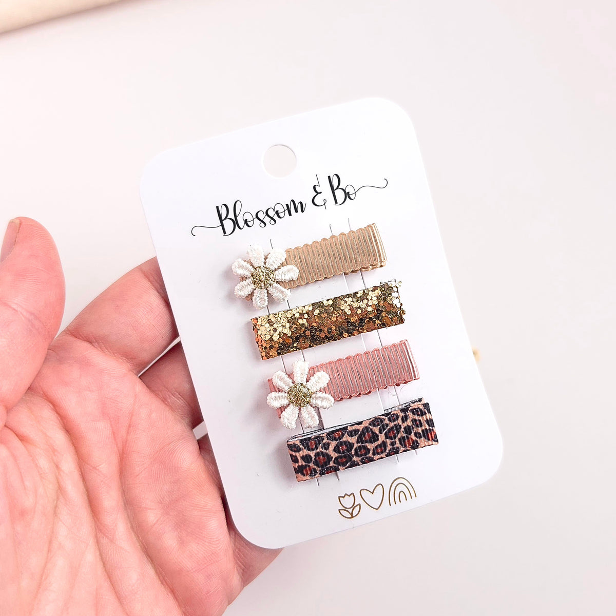 A hand holding a white card displaying four hair clips. The card is branded 'Blossom & Bo' and features four clips: a beige ribbon clip, a gold glitter clip with a white flower accent, a dusty pink ribbon clip, and a leopard print clip.