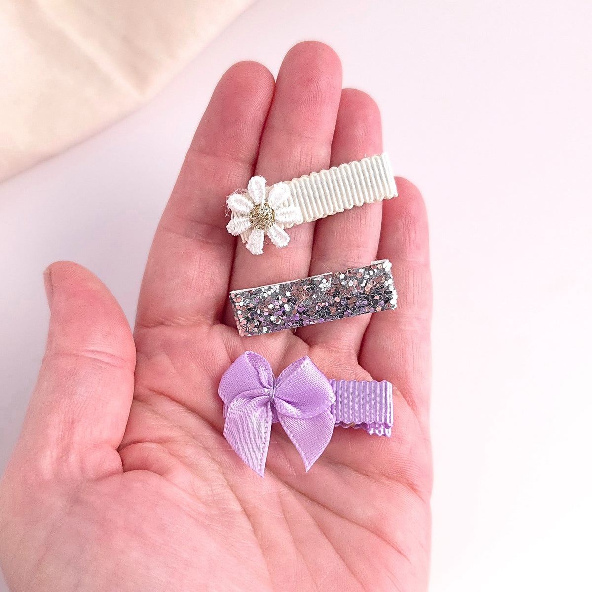 Three small hair clips held in a person's hand: a white fabric flower clip with a gold center, a silver glitter bar clip, and a lavender satin bow clip.