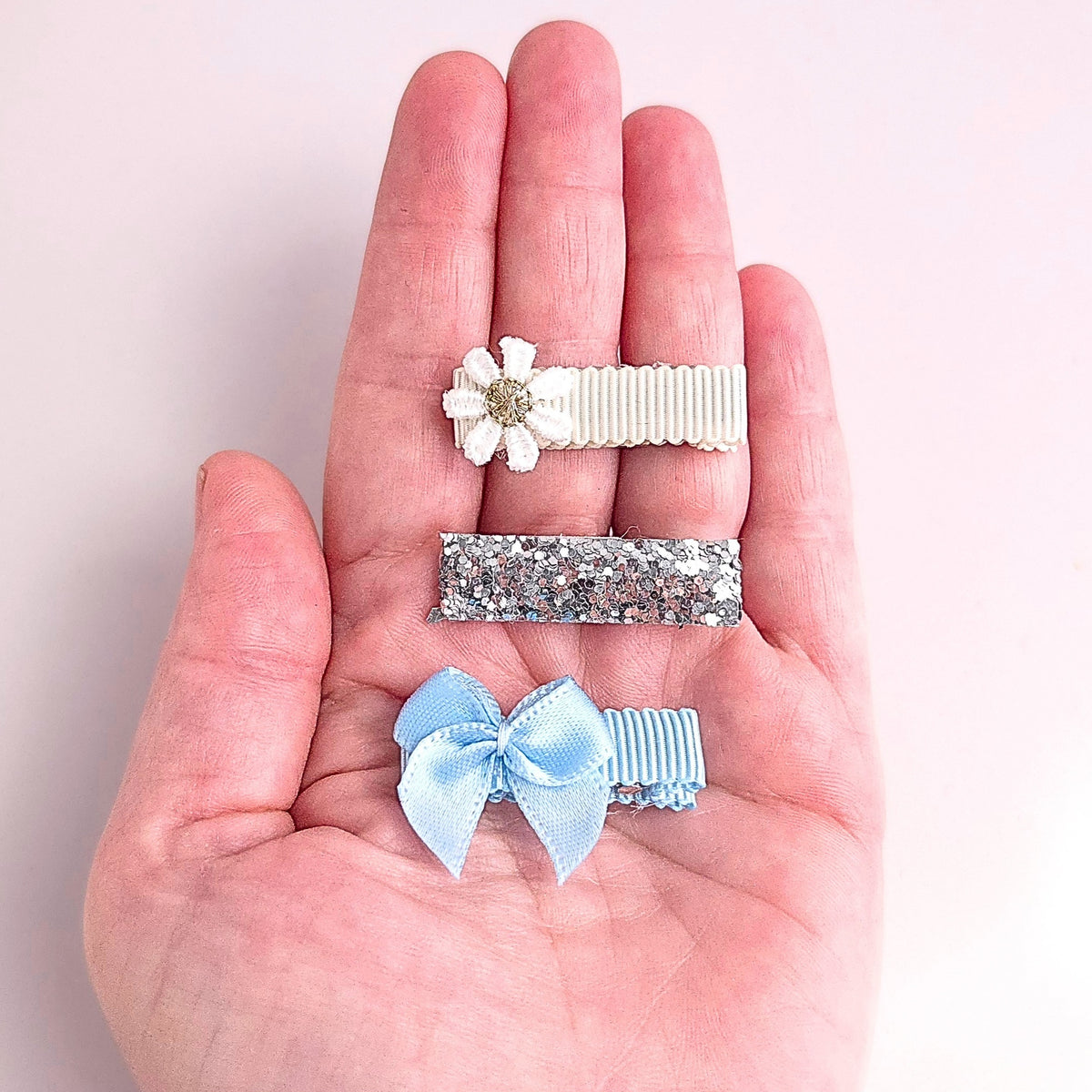 Baby and toddler hair clips shown in hand, with white daisy, silver glitter and blue bow