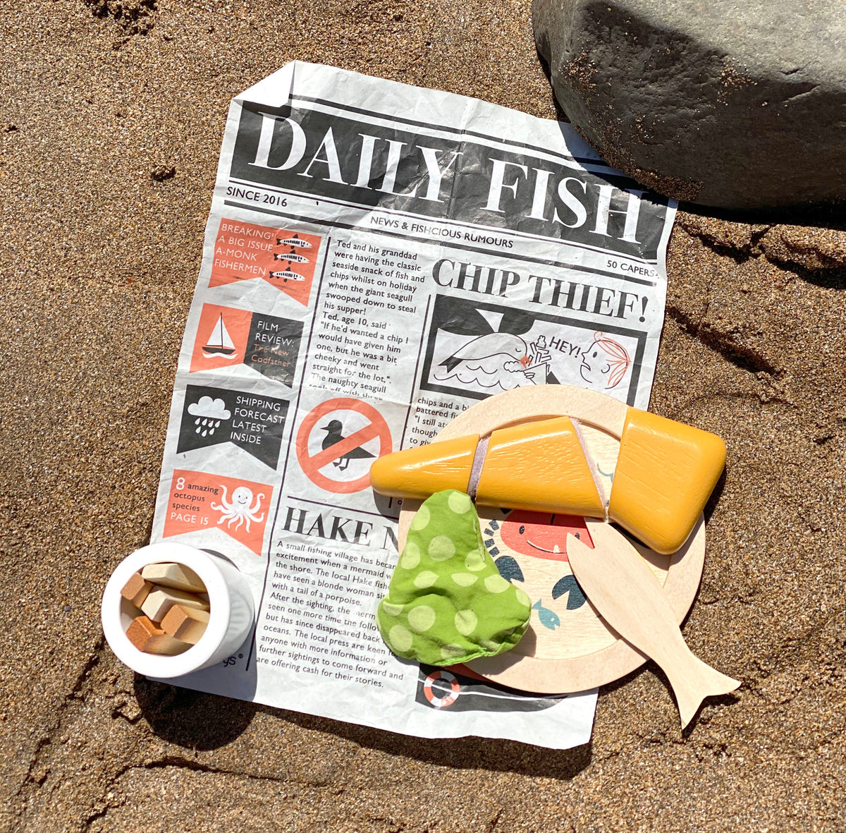 Toy fish, newspaper, and cup on a sandy surface