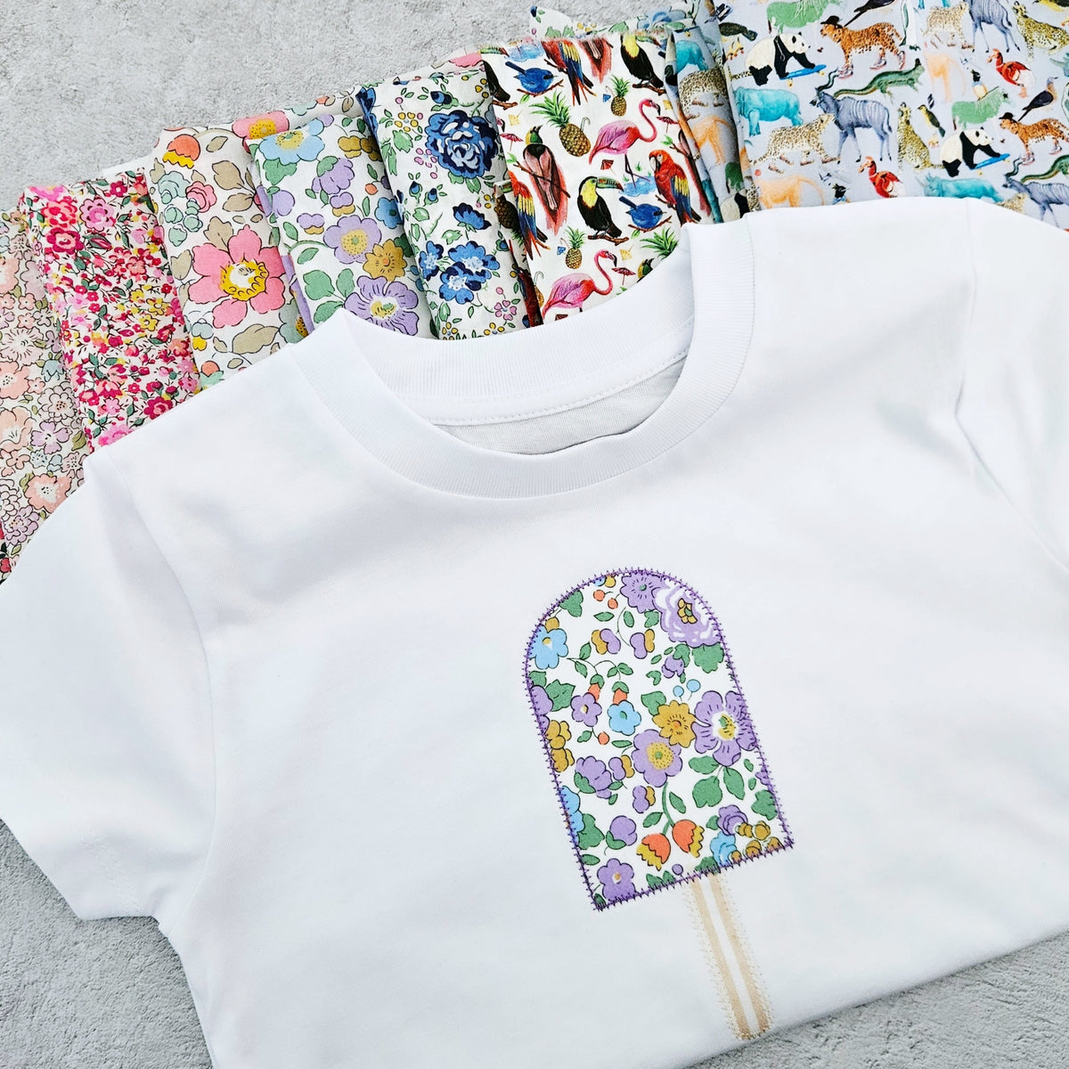 Close up of a children's white t-shirt with a fabric Liberty of London ice lolly applique on the front. Samples of fabrics behind.