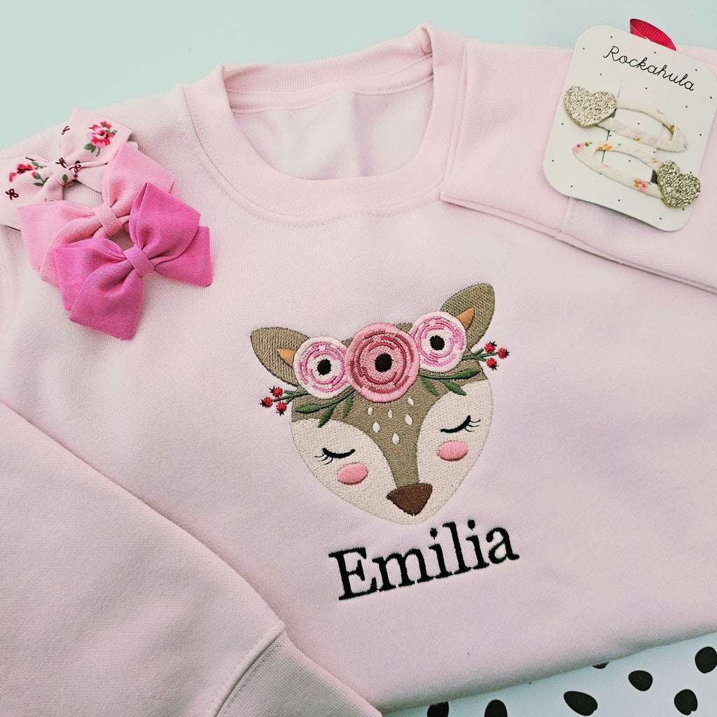 A light pink crewneck sweatshirt featuring an embroidered fox face with a pink floral crown and the name 'Emilia' in black script. The sweatshirt is styled with a pink fabric bow and a card of matching hair clips on a pale blue background.