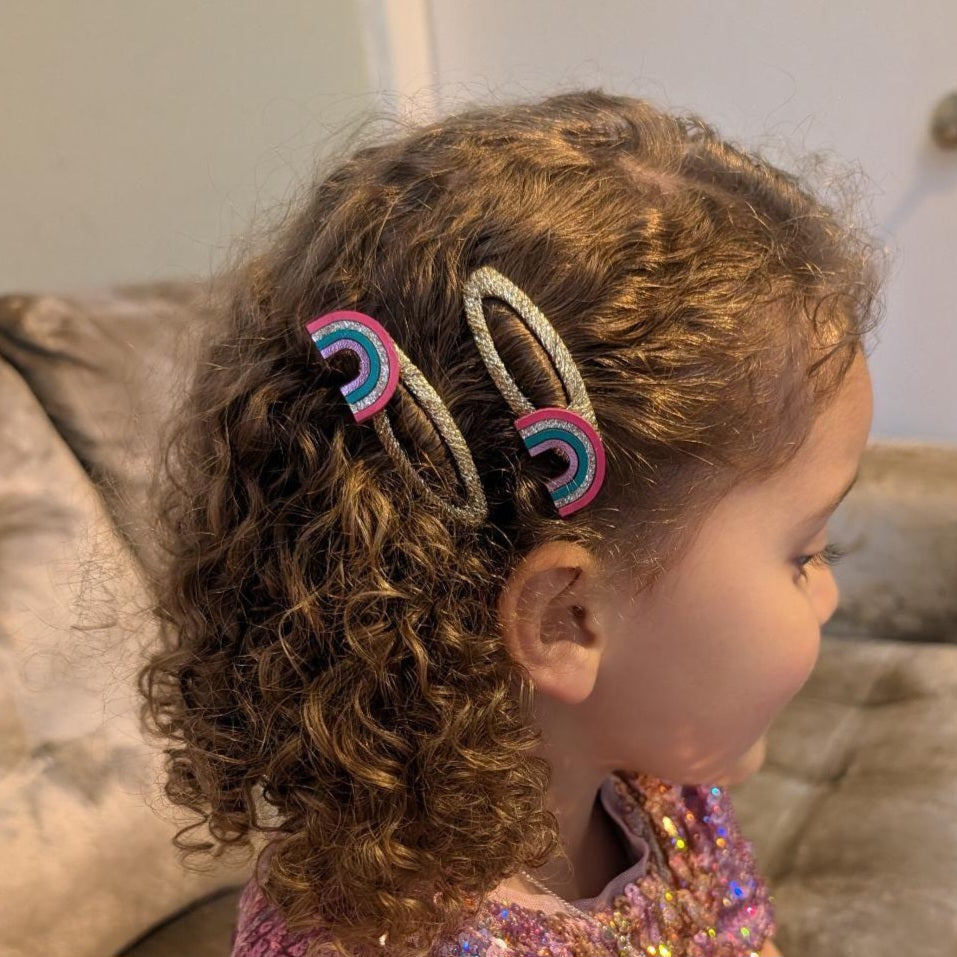 A close-up side view of a young child with curly brown hair wearing two rainbow-patterned hair clips, set against a soft-focus indoor background.