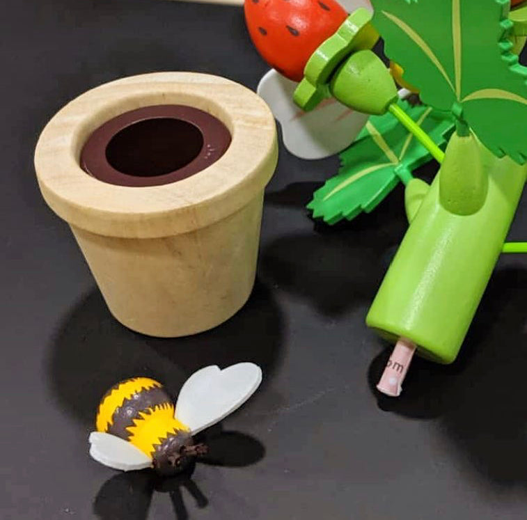 Wooden flower pot with a bee toy and a strawberry plant toy figure on a dark surface