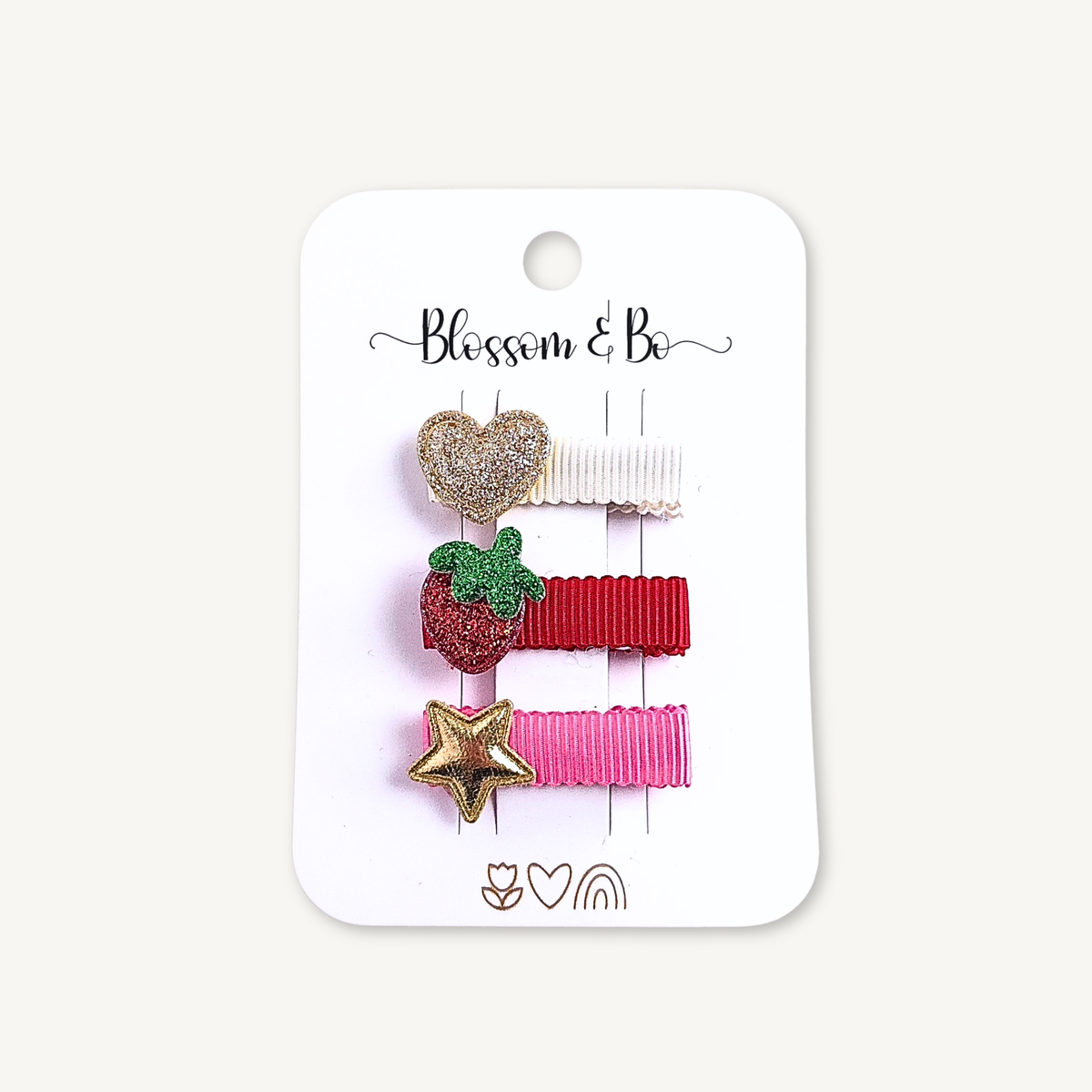 A set of three small hair clips on a white display card. The clips include a glittery gold heart, a red strawberry with green leaves, and a gold star, attached to three ribbons in cream, red, and pink.