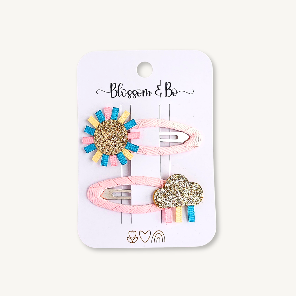 Set of two fabric covered snap hair clips featuring a glitter sun and rainbow cloud on a Blossom & Bo display card.