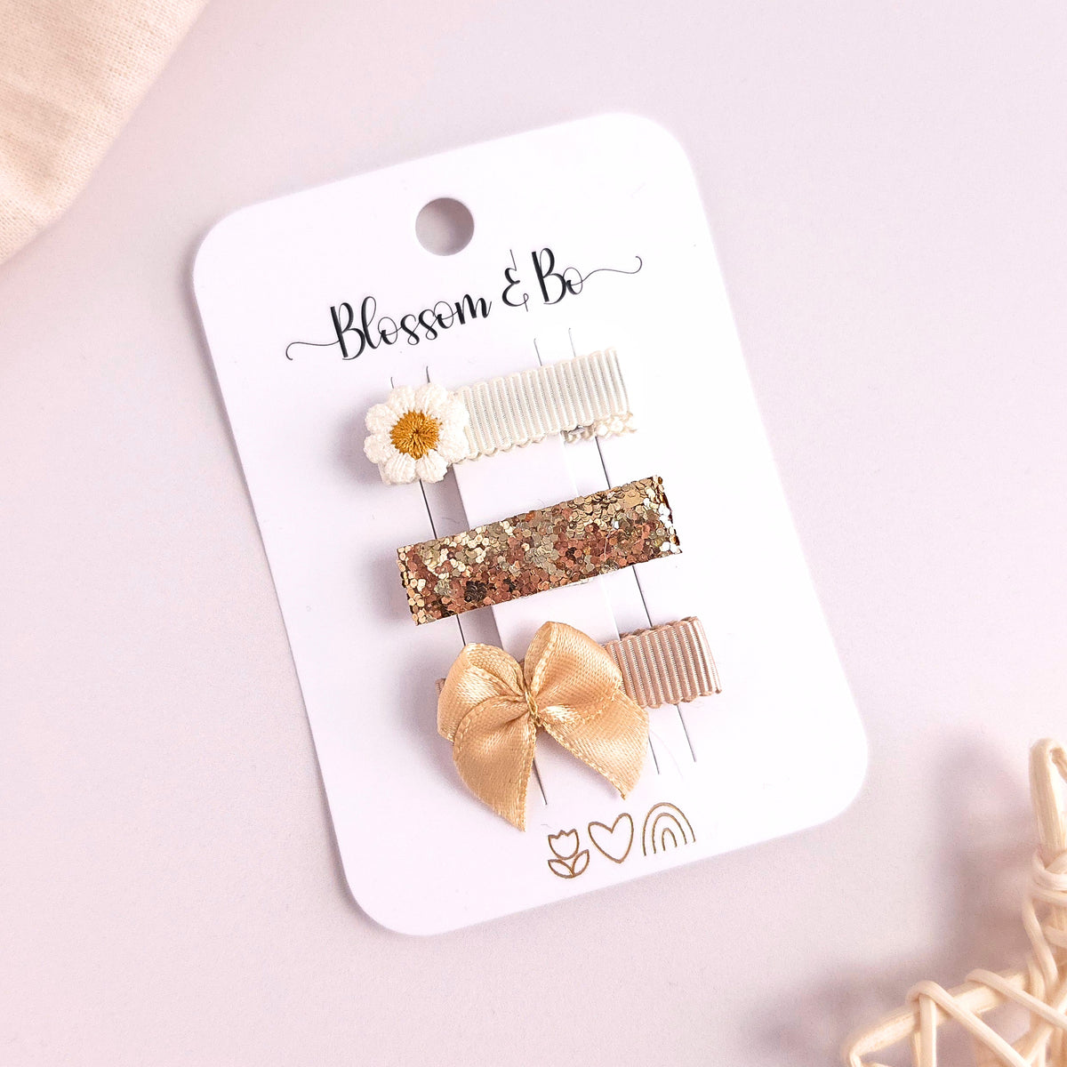 Three hair clips attached to a white card labeled 'Blossom & Be,' photographed on a pale pink background. The clips include a white daisy, a gold glitter bar, a beige ribbed clip, and a gold satin bow.
