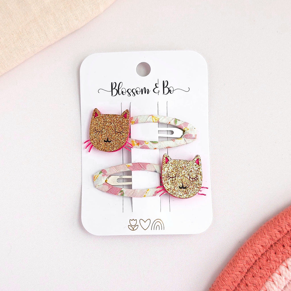 Two hair clips with glittery cat faces and pastel floral bands, displayed on a white card labeled 'Blossom & Bo' on a light pink surface.