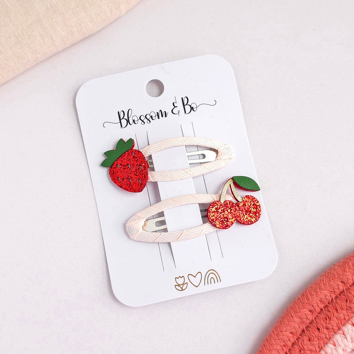 Two cream-colored hair clips decorated with a red glittery strawberry and a pair of red glittery cherries with green leaves, attached to a white card printed with 'Blossom & Be'.
