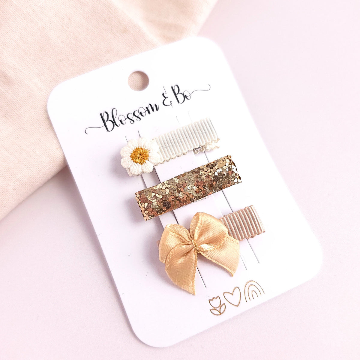 Three hair clips on a white display card labeled 'Blossom & Be,' photographed on a soft pink background. The clips include a white daisy with gold center, a white lace comb, a gold glitter bar, and a gold satin bow.