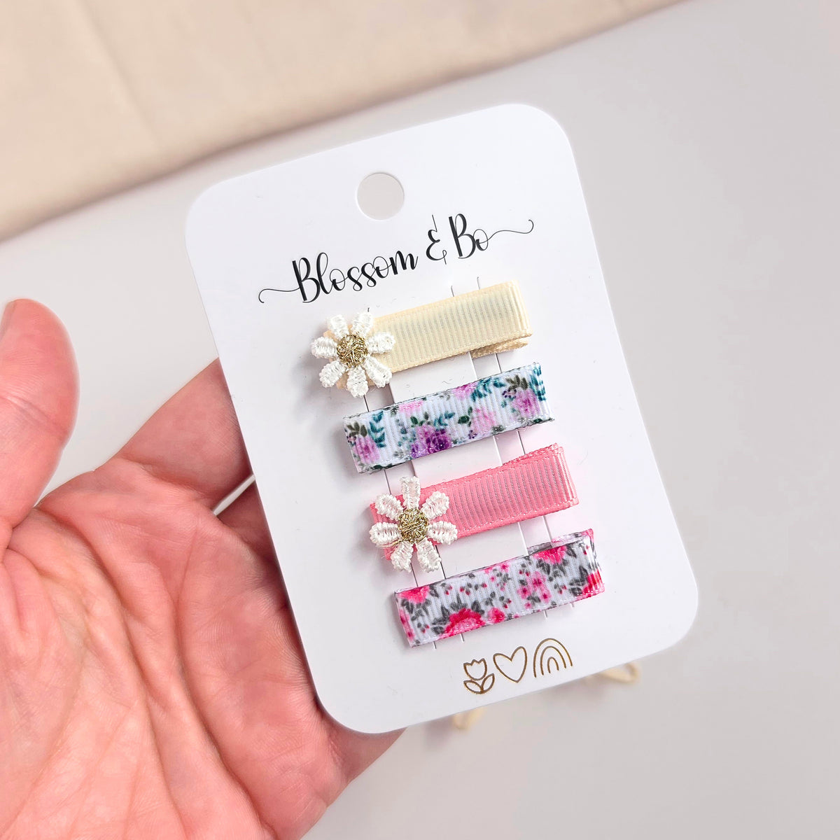Four hair clips on a white display card labeled 'Blossom & Bo', held in a hand. The clips include floral ribbon styles and two cream flower clips with gold centers.