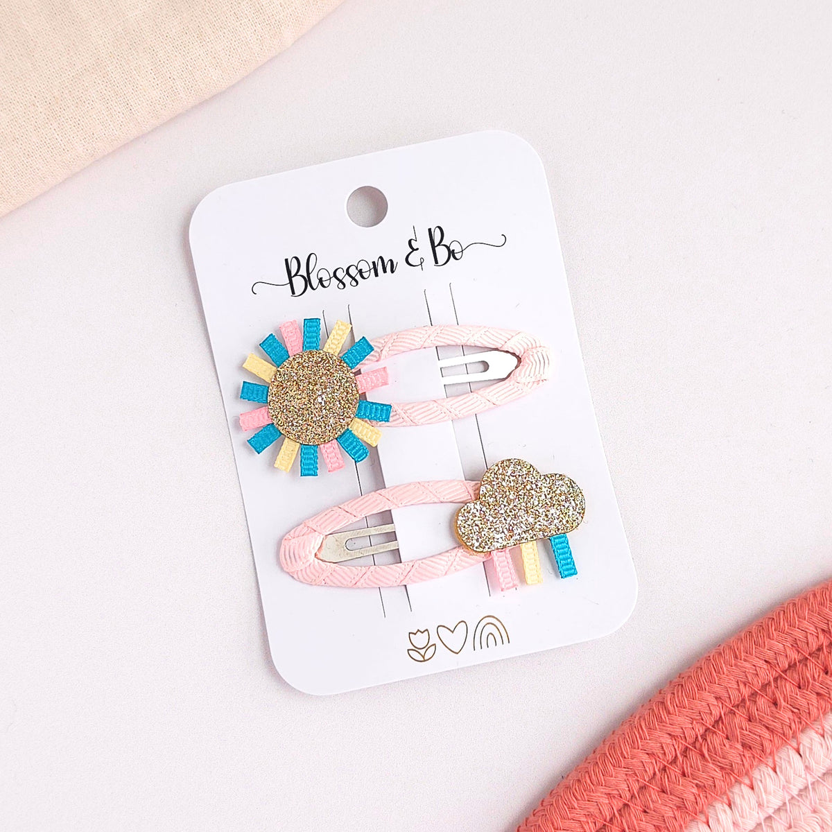 Two pastel hair clips on a white card labeled 'Blossom & Bo'. One clip has a sunburst design with a glittery gold center and blue, pink, and yellow ribbon rays.