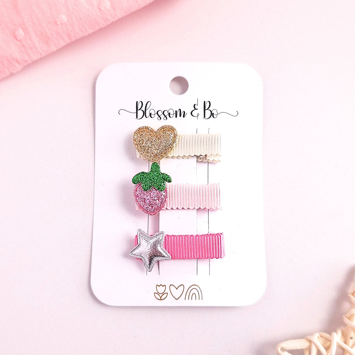 A set of three pastel hair clips on a white display card. The clips include a glittery gold heart, a pink glitter strawberry with green leaves, a pale pink ribbon clip, and a silver glitter star on a pink ribbon clip.