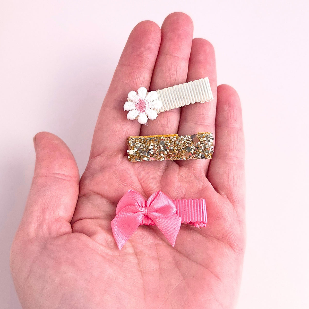 Three small hair clips displayed on an open hand against a pale pink background: a white flower clip with a pink center, a glittery gold clip, and a pink ribbon bow clip.