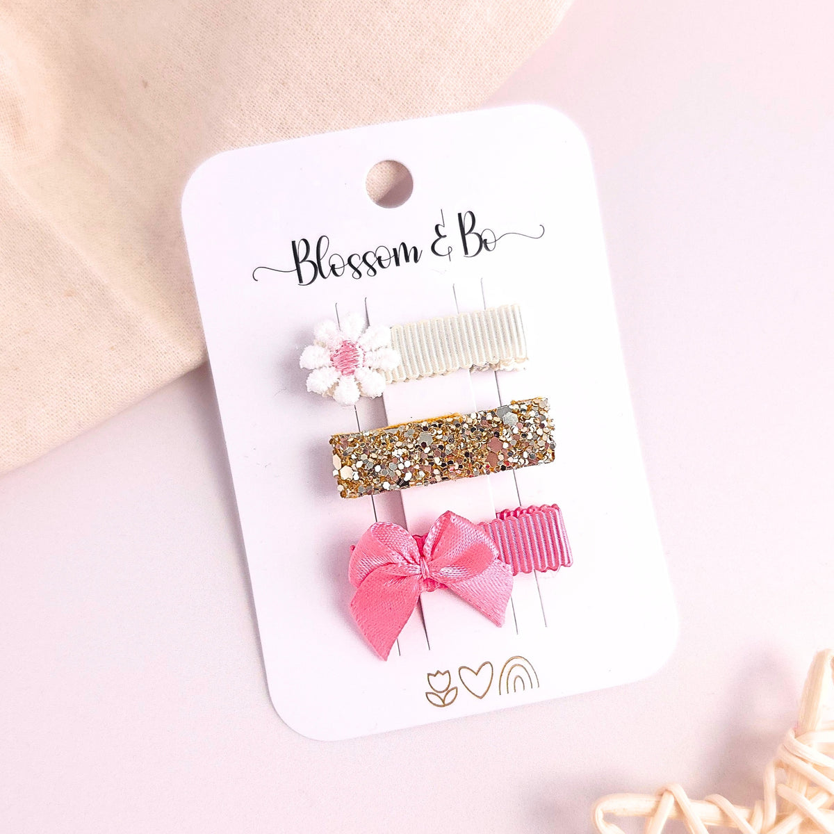 Three hair clips on a white card labeled 'Blossom & Bo' against a soft pink background with decorative fabric props.