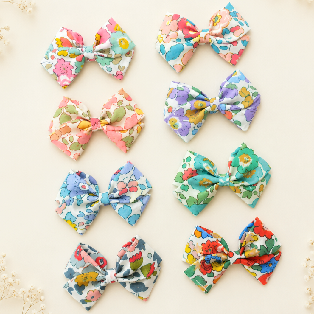 Eight fabric hair bows arranged in a grid on a light cream background. Each bow features a colorful floral print with various shades of blue, pink, red, yellow, and green on white bases.
