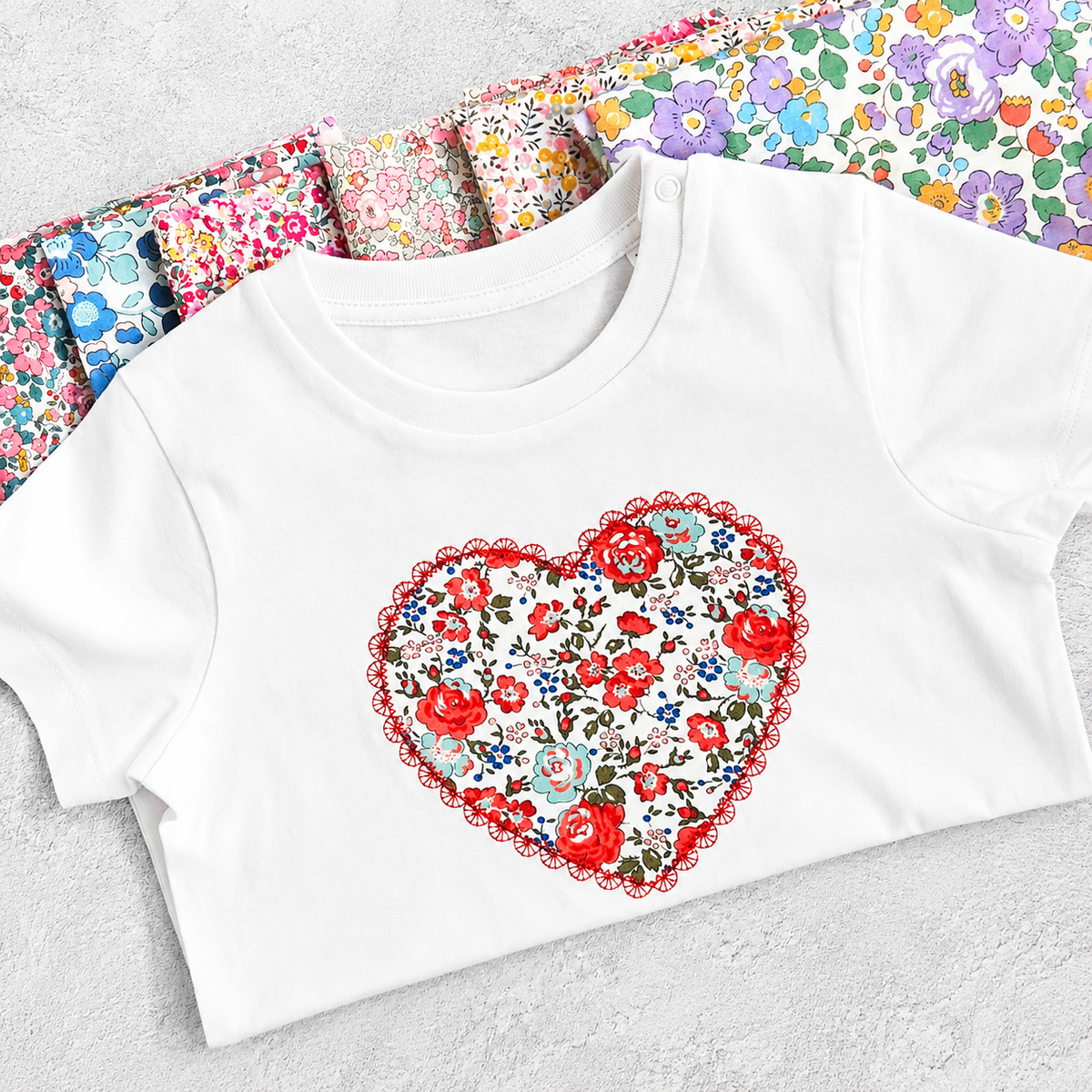 A white t-shrt with red floral heart on the front, folded and sitting on different fabric choices.