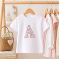 A white short-sleeve children's T-shirt hanging on a wooden hanger, featuring a large decorative capital letter 'A' on the chest filled with a pink floral pattern. The shirt is displayed on a wooden clothing rack alongside other pastel-colored baby clothes, with a wicker basket and books in the background.