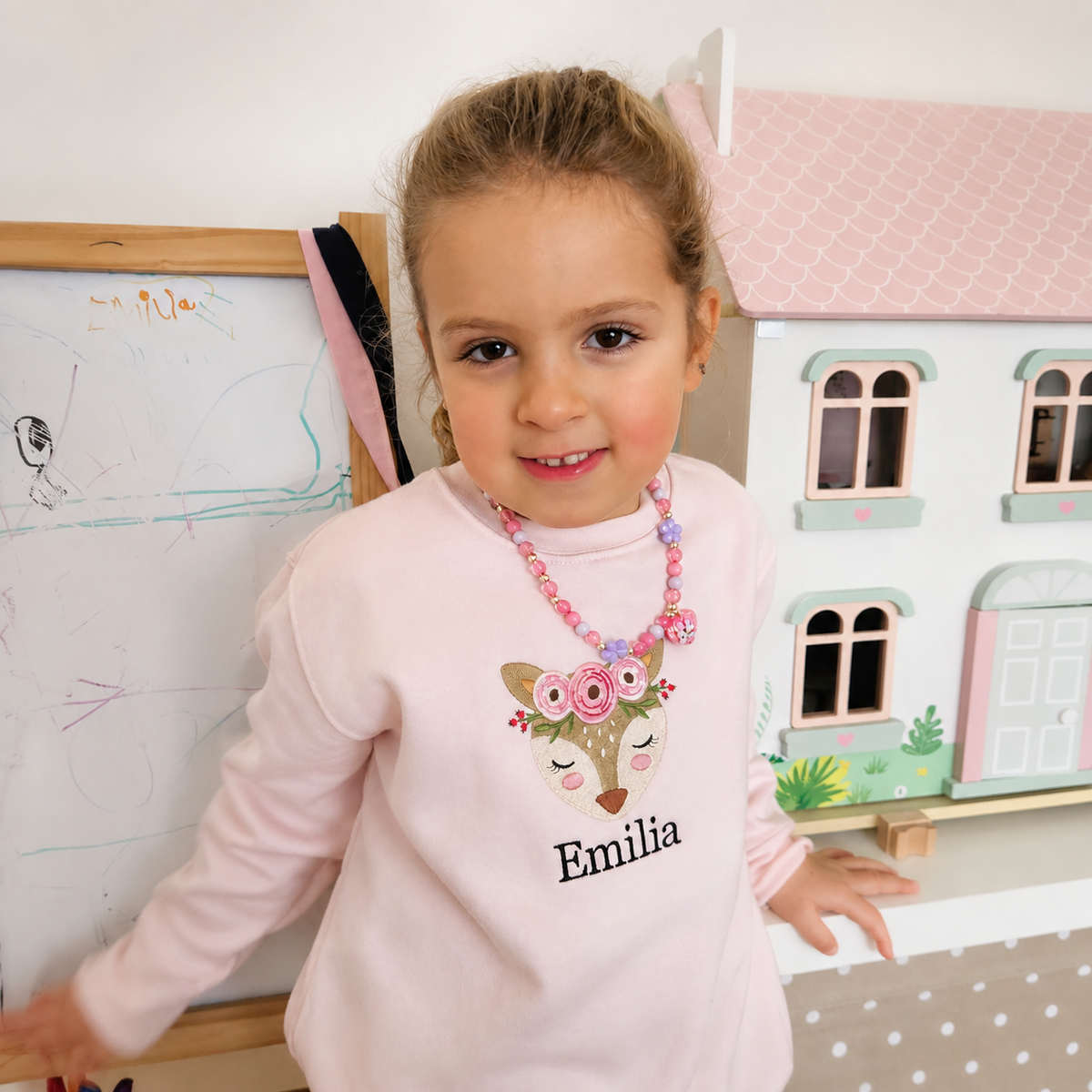 A young girl with blonde hair in a ponytail stands indoors wearing a light pink long-sleeve sweatshirt with a personalized deer graphic and the name 'Emilia' printed on the front. She is also wearing a pink beaded necklace.