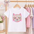 A white short-sleeve kids' t-shirt hanging on a wooden hanger, featuring a colorful floral cat face graphic on the front. The shirt is displayed on a clothing rack alongside other pastel-colored garments, with a decorative bow on the wooden hanger and a vase of flowers in the background.