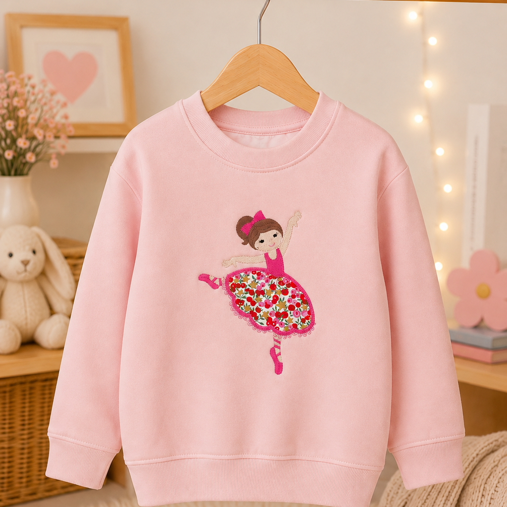A light pink crewneck sweatshirt hanging on a wooden hanger, featuring a graphic of a ballerina with a colorful floral skirt. The sweatshirt is displayed on a wooden clothing rack in a cozy, softly lit room with a plush bunny, framed heart art, and warm fairy lights in the background.