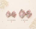 Two floral fabric hair bows shown on a light cream background with decorative flowers and confetti. One bow is labelled 2.5 inches and one is 3 inches.