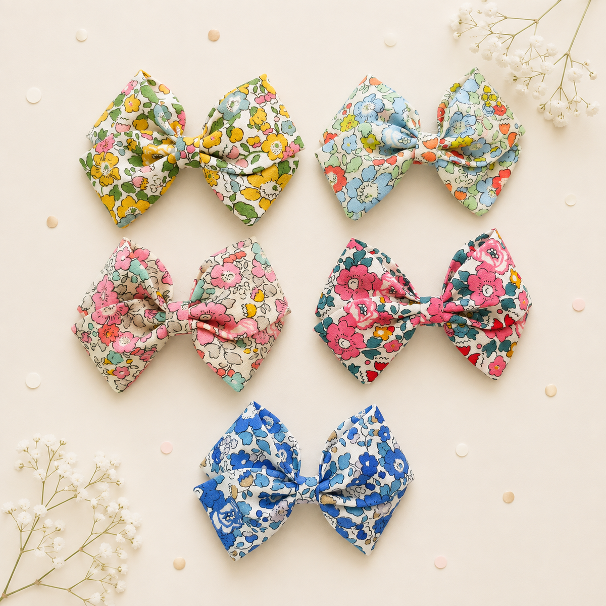 Five fabric hair bows arranged on a light beige background with small white flowers and scattered confetti. The bows feature different floral prints in blue, pink, yellow, green, and red on white or pastel bases.