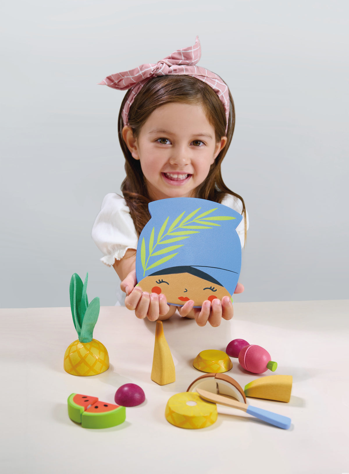 Child holding a colorful toy head with a blue hat and green leaf, surrounded by various toy fruits and vegetables on a light gray background.
